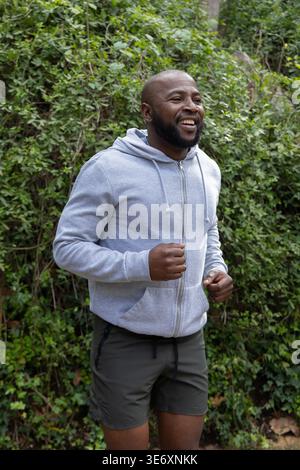 African American man jogging in park by dense ivy wearing light-gray zip-up hoodie and dark shorts Stock Photo