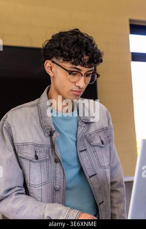 Adult male wearing denim jacket and round eyeglasses leaning at desk in office, working on laptop Stock Photo