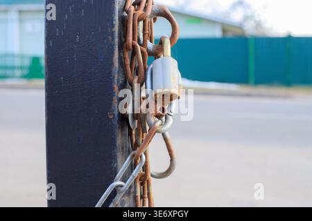 Close-up of weathered rusty chains and a padlock on a black metal pillar. Industrial security locking system for restricted urban areas, construction Stock Photo