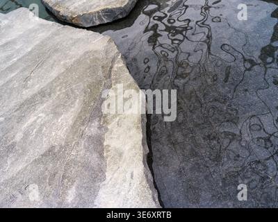 High angle close-up view of rough, light-colored natural stone slabs forming the edge of a calm, dark water pool that reflects abstract wavy patterns Stock Photo