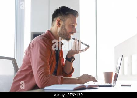 Man Using Voice Command on Laptop While Sitting at a Desk in a Bright Room Stock Photo