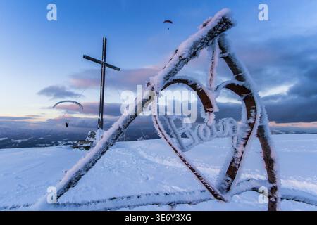 View of a snow-covered cross and heart-shaped structure with paragliders soaring in the distance, against a twilight sky, Schöckl, Styria, Austria. Stock Photo