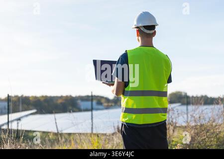 A young male electrical engineer checks solar panels while using a laptop in a renewable energy area. Stock Photo