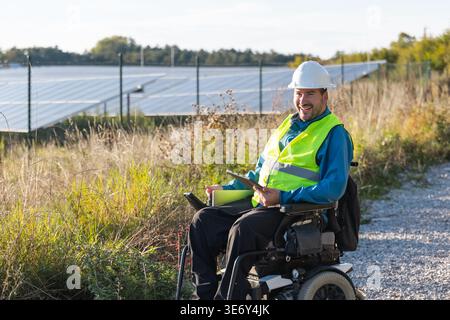 A male electrical engineer portrait in a wheelchair working and using a laptop near solar panels in a renewable energy area. Stock Photo