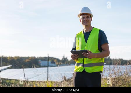 A young male electrical engineer inspects solar panel installations while using a tablet at a renewable energy location in daylight. Stock Photo
