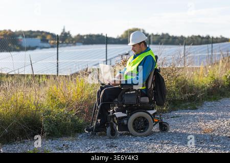 Male engineer in a wheelchair uses a laptop to check solar panel systems in a renewable energy field under clear skies. Stock Photo
