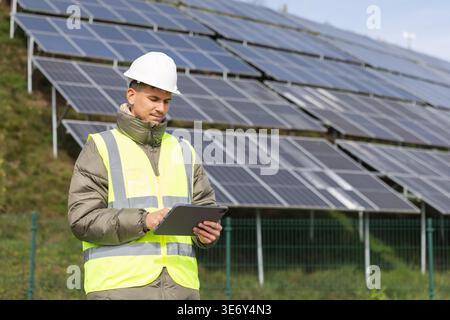 Young male electrical engineer uses a tablet to inspect solar panel installations in a renewable energy area during daylight hours. Stock Photo