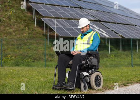 Male engineer in wheelchair uses laptop to work on solar panel systems in the renewable energy field during daylight. Stock Photo