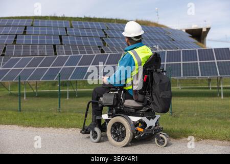 A male electrical engineer in a wheelchair uses a laptop while working with solar panels in a renewable energy environment. Stock Photo