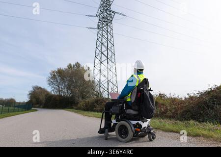 A male electrical engineer with disability in a wheelchair uses a laptop to monitor power lines outdoors while wearing safety gear. Stock Photo