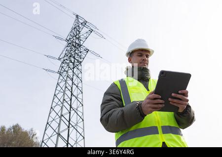 Young male engineer uses a tablet to check high-voltage power lines while working outdoors in energy infrastructure. Stock Photo