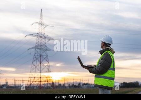 A young male electrical engineer is checking high-voltage power lines with a laptop outdoors at sunset. He wears a safety helmet and vest. Stock Photo