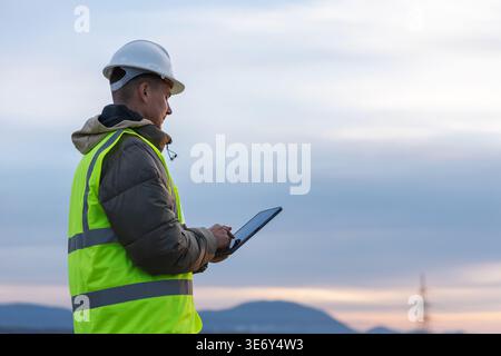 A young male electrical engineer works outdoors, inspecting high-voltage power lines with a tablet in a utility area at dusk. Stock Photo