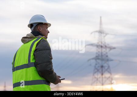 Young male electrical engineer checks high-voltage power lines using a laptop in an outdoor area during sunset hours. Stock Photo