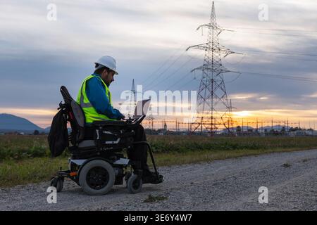 A male electrical engineer with disability in a wheelchair uses a laptop to check high-voltage transmission lines during sunset. Stock Photo