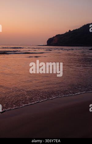 View of a tranquil beach where the gentle waves kiss the shore under a sky painted with soft hues of pink and orange at dusk, Chiplun, Maharashtra, In Stock Photo