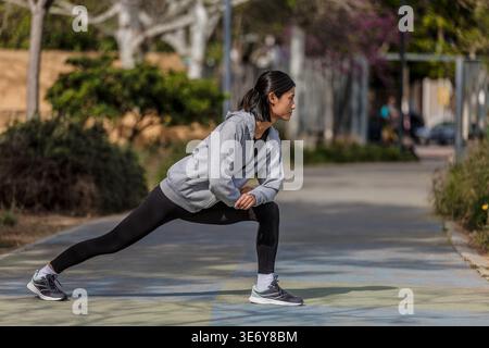 A woman of Asian descent stands in a park, performing a stretching exercise. It is a sunny day, and she is focused on warming up for her workout. The Stock Photo