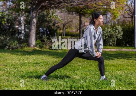 A young Asian woman stretches her body in a park in Spain. She practices outdoor exercises on grass under clear skies during daytime. This activity pr Stock Photo