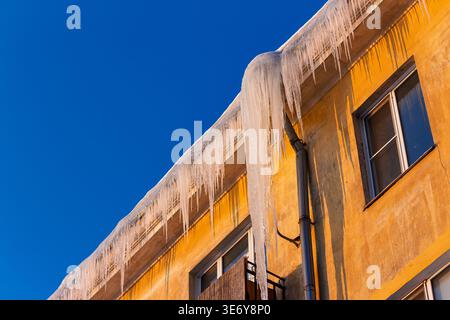 Long, sharp icicles hang from a sun-kissed old facade, reflecting the last rays of golden light under a stark blue winter sky Stock Photo