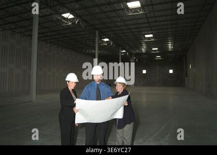 Corporate representatives looking at blueprints with construction engineer inside new empty warehouse, New Jersey USA Stock Photo