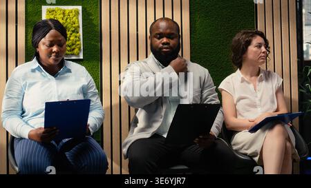 Multiethnic job seekers waiting in the corridor with their CV files, preparing for formal interview with the HR. Applicants reflecting impatience and confidence during head hunting. Stock Photo