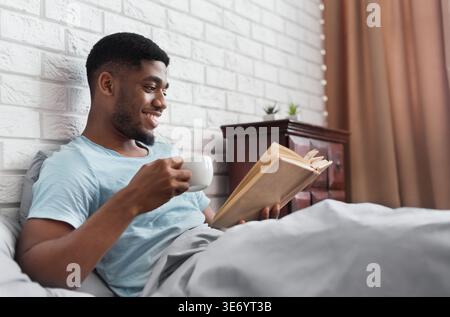 Black man drinking coffee and reading book in bed Stock Photo
