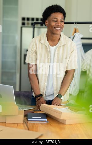 Man packing cardboard boxes on wooden table with laptop and clothing rack in small home studio Stock Photo