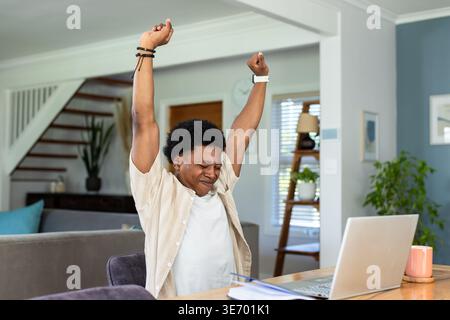 African American man celebrating, raising arms at wooden table with open laptop, papers and mug Stock Photo