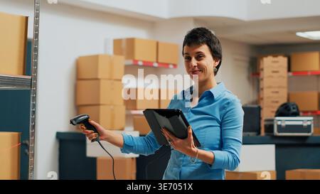 Young employee scanning products from warehouse, working with scanner to do inventory and check supply chain logistics. Female entrepreneur using tablet to scan merchandise bar code. Handheld shot. Stock Photo