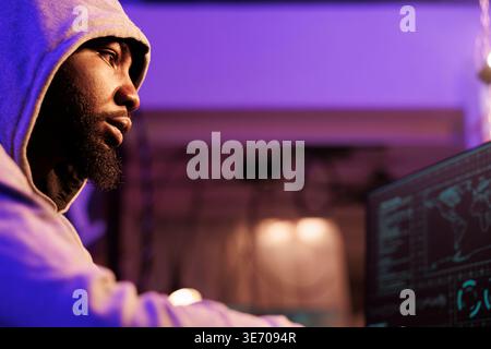 African american hacker using malware to access server in abandoned warehouse. Young criminal in hood using spyware and planning internet phishing attack in dark room hideout Stock Photo