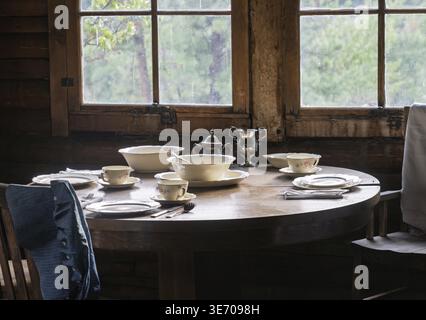Rustic cabin interior with a round wooden table set with elegant tableware, including bowls, tea set, and plates, by the window Stock Photo