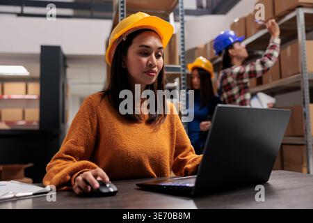 Warehouse manager working on automated inventory management on laptop while sitting at desk in storage room. Storehouse operator in yellow hardhat analyzing customer orders data on computer Stock Photo