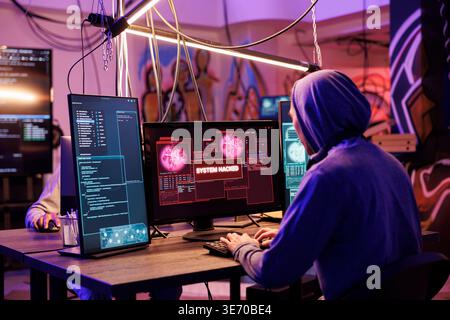 Criminal in hood successfully hacking into computer system in abandoned warehouse. Hacker gaining unauthorized access and stealing valuable data from database online server Stock Photo