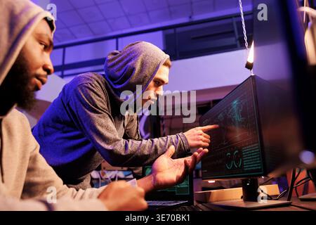 Hackers pointing at map location at computer screen while spying online. Two diverse young men in hoods using illegal spyware while doing criminal activity together at night Stock Photo