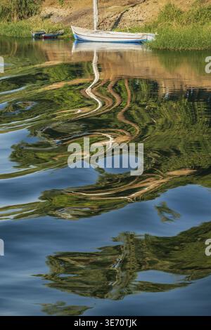 A small sailboat floats on a calm river Nile, its reflection distorted by gentle ripples creating an abstract pattern on the water's surface Stock Photo