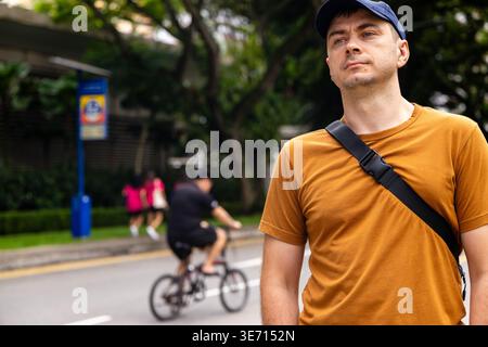 Man wearing blue cap and orange t-shirt stands on urban sidewalk looking away. Blurred cyclist and pedestrians pass by on city street background. Auth Stock Photo