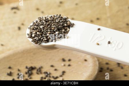 Superfood known as black chia seeds in white plastic measuring spoon over wood bread board on kitchen table Stock Photo