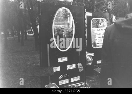 Slaviansk, 1980s, Lenin Culture and Recreation Park (now Shevkovychny Park). An outdoor exhibition or trade fair of Soviet consumer goods displayed in the park. Several dark stands feature various items for sale: large framed oval mirrors, kitchen utensils like spatulas and scoops, a portable grill with skewers, and other household appliances. The mirrors reflect different passersby, including a boy eating something, and other distant stalls. Trees form the background of this public space. Peaceful Donbas life before the conflict. Stock Photo