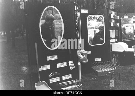 Slaviansk, 1980s, Lenin Culture and Recreation Park (now Shevkovychny Park). An outdoor exhibition or trade fair of Soviet consumer goods displayed in the park. Several dark stands feature various items for sale: large framed oval mirrors, kitchen utensils like spatulas and scoops, a portable grill with skewers, and other household appliances. The mirrors reflect different passersby, including a boy eating something, and other distant stalls. Trees form the background of this public space. Peaceful Donbas life before the conflict. Stock Photo