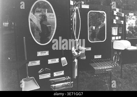 Slaviansk, 1980s, Lenin Culture and Recreation Park (now Shevkovychny Park). An outdoor exhibition or trade fair of Soviet consumer goods displayed in the park. Several dark stands feature various items for sale: large framed oval mirrors, kitchen utensils like spatulas and scoops, a portable grill with skewers, and other household appliances. The mirrors reflect different passersby, including a boy eating something, and other distant stalls. Trees form the background of this public space. Peaceful Donbas life before the conflict. Stock Photo