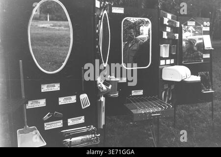 Slaviansk, 1980s, Lenin Culture and Recreation Park (now Shevkovychny Park). An outdoor exhibition or trade fair of Soviet consumer goods displayed in the park. Several dark stands feature various items for sale: large framed oval mirrors, kitchen utensils like spatulas and scoops, a portable grill with skewers, and other household appliances. The mirrors reflect different passersby, including a boy eating something, and other distant stalls. Trees form the background of this public space. Peaceful Donbas life before the conflict. Stock Photo