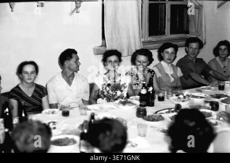 Slaviansk, 1959, Energy Technical College. A close-knit group of graduates and guests sits at a festive dining table to celebrate the inaugural graduation class. In the center, a smiling woman proudly holds a large, beautiful bouquet of wildflowers. The table is set with various plates of food, glasses, and dark bottles for the celebratory feast. The attendees wear formal mid-century attire, capturing a wonderful moment of pride and camaraderie in a warmly lit room. Peaceful Donbas life before the conflict. Stock Photo