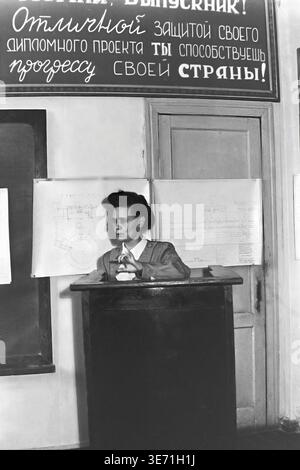 Slaviansk, 1959, Energy Technical College. A young female graduate confidently defends her diploma project at a wooden podium. Behind her, engineering blueprints are pinned to a door. A prominent propaganda banner hangs above, reading 'With an excellent defense of your diploma project, you contribute to the progress of your country!' in Cyrillic. Dressed in a formal mid-century outfit, the young specialist perfectly represents the dedicated and hard-working post-war youth. Peaceful Donbas life before the conflict. Stock Photo