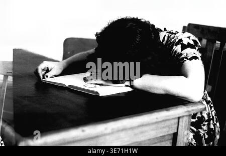 Sloviansk, 1959, Energy Technical College. A young female student rests her head on an open book while sitting at a wooden desk, seemingly exhausted from rigorous studying. This candid black and white archival photograph beautifully captures the intense dedication and academic effort of youth during the Soviet educational era. The intimate interior scene highlights the everyday reality and intense educational pursuits of the first graduating class, reflecting a moment of quiet, well-earned rest in a peaceful Donbas. Stock Photo