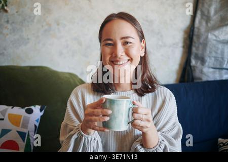 Wellness, portrait or Asian woman on sofa with coffee, positive attitude or chill on weekend break. Happiness, peaceful or person in house with warm Stock Photo