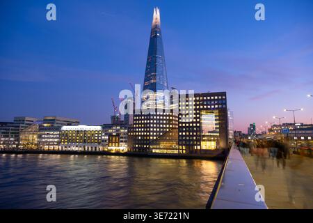 The Shard stands tall against the evening sky in London. People walk along the riverbank as lights from buildings reflect on the water. Construction cranes can be seen nearby. Stock Photo