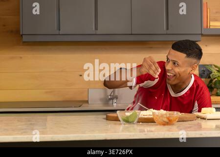 Man preparing meal on stone countertop wearing red athletic shirt, chopping cheese with lime wedges. Kitchen, cooking, smile, home, counter, salsa, pl Stock Photo