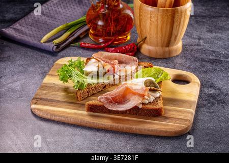 A rustic still life of two open-faced sandwiches on a wooden cutting board. Toasts feature rye bread, prosciutto, soft cheese, and lettuce on a dark, Stock Photo