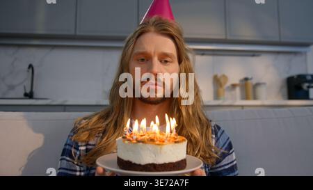 Sad lonely Caucasian man sitting on couch at home holding birthday cake party alone upset depressed male wearing festive hat feeling isolated Stock Photo