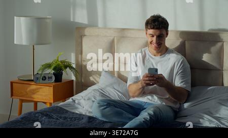 Smiling Caucasian man lying bed at home bedroom using smartphone online chatting carefree male joyful guy laughing browsing internet social media Stock Photo